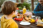 Father dad school kid boy child having a picnic in the forest camping site with vegetables, juice, coffee, and croissants. Wooden crate with fresh organic veggies surrounded with bread baguettes.