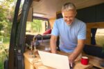 Senior Man On Camping Trip In Countryside Working Inside RV Using Laptop