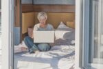 Senior smiling woman with short gray hair, using laptop on bed. At country home, in hotel,in camping