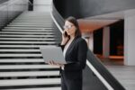 The Environmental Benefits Of Remote Work business woman in a formal suit with a laptop in the office of the business center stands on the steps in the lobby and works online