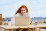 Freelancer woman sitting in cafe on street and remotely working on laptop. Female student listening courses