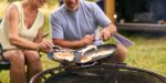 Close Up Of Senior Couple Camping In RV Cooking Bacon And Eggs For Breakfast Outdoors On Fire