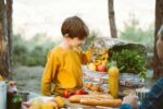 Father dad school kid boy child having a picnic in the forest camping site with vegetables, juice, coffee, and croissants. Wooden crate with fresh organic veggies surrounded with bread baguettes.