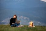 young woman freelancer with laptop in the mountains in the evening. Tourist girl sitting near campfire