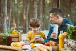 Father dad and school kid boy child having a picnic in the forest camping site with vegetables, juice, coffee, and croissants. Wooden crate with fresh organic veggies surrounded with bread baguettes.